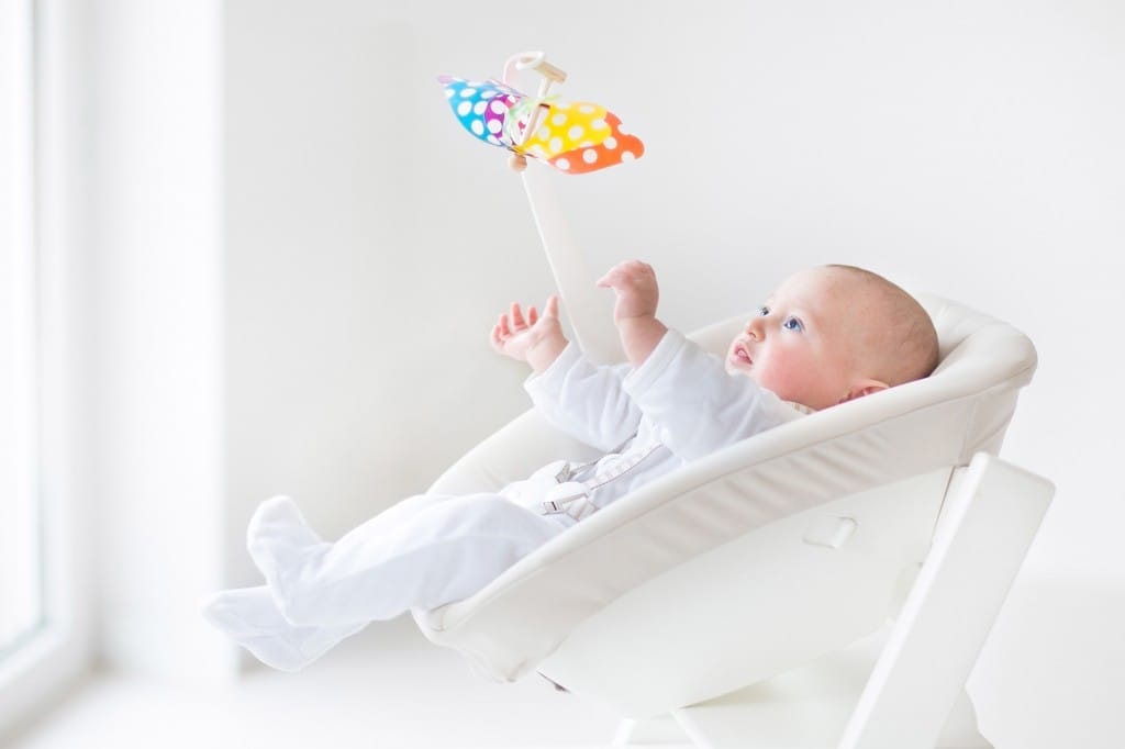 Cute newborn baby boy watching a colorful mobile toy sitting in a white chair next to a window